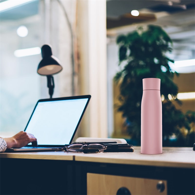Person using a laptop on a desk with a pink water bottle and glasses nearby, blurred office background